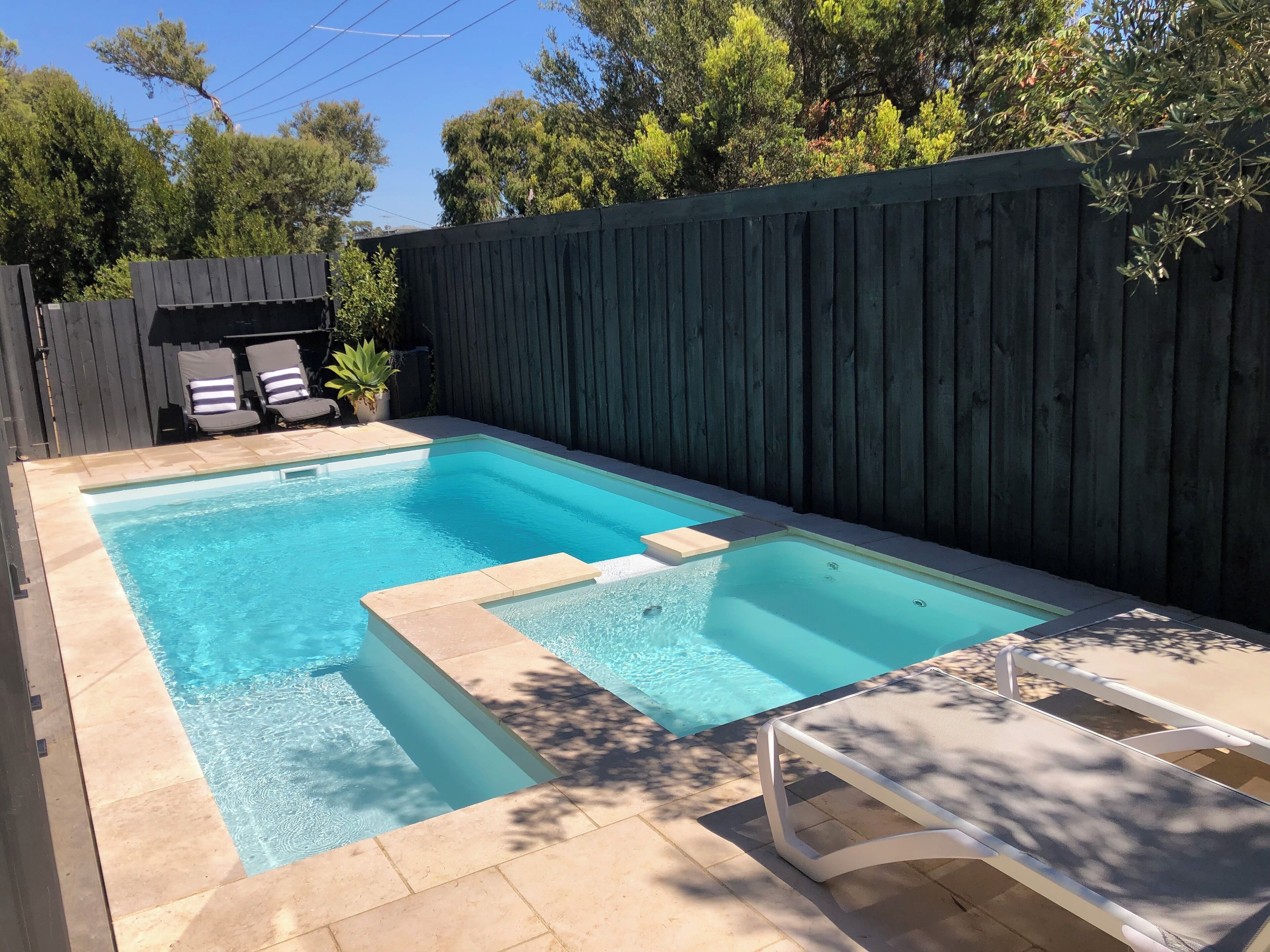 A rectangular swimming pool with a tiled deck, surrounded by a wooden fence and trees.