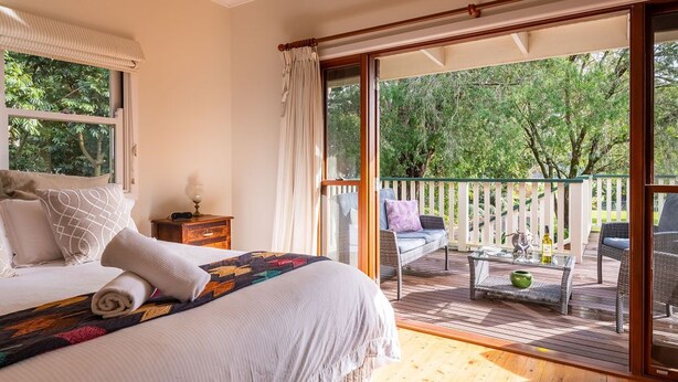Une chambre avec un grand lit, une table de chevet et une vue sur une terrasse en bois et des arbres.