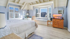 A bedroom with a large bed, a wooden dresser, a television, and a window with a view of a building. - Gated Fairytale Estate | Lakefront w/ Ocean Views (Oceanside)