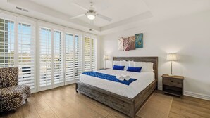 A bedroom with a large bed, a ceiling fan, a nightstand, and a view through a window with blinds. - Seaside Delight | 350 ft from the beach | Community Pool (Virginia Beach)