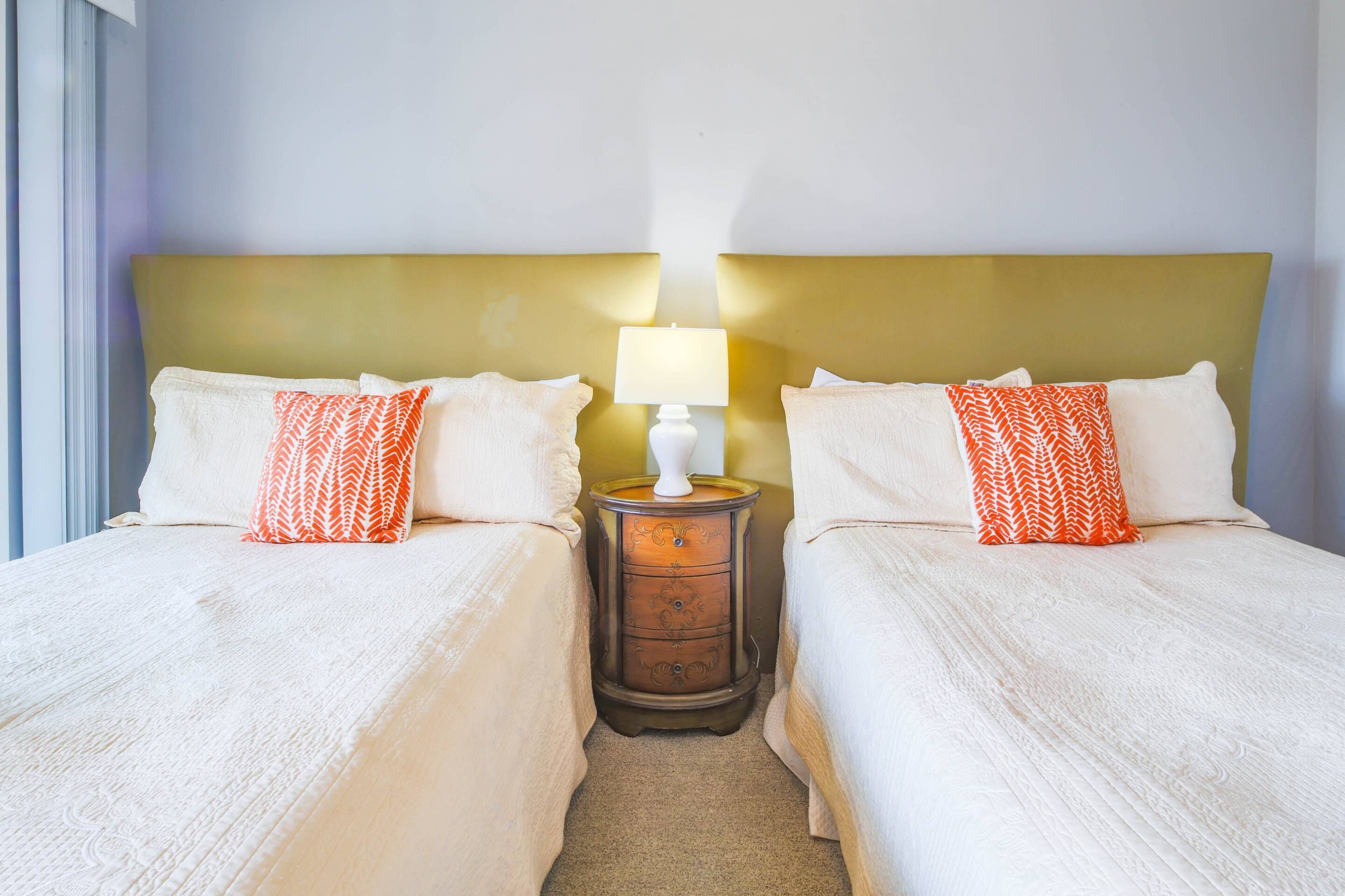 Quarto de hotel com duas camas, cada uma com roupa de cama branca e almofadas laranja estampadas. Há uma mesa de cabeceira com uma luminária entre as camas.