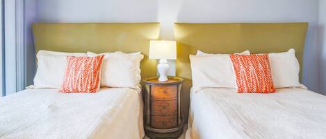 Quarto de hotel com duas camas, cada uma com roupa de cama branca e almofadas laranja estampadas. Há uma mesa de cabeceira com uma luminária entre as camas.