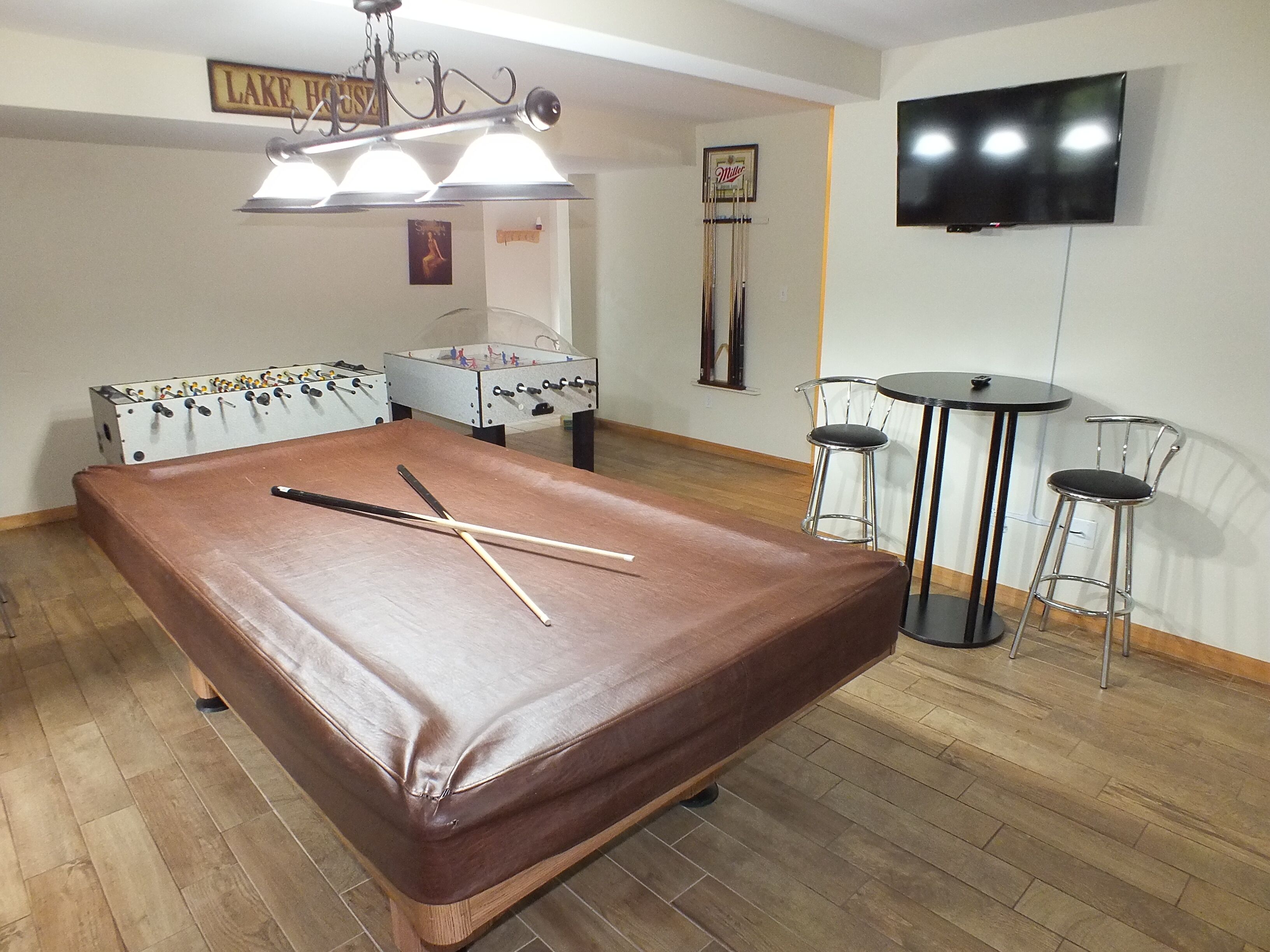 A room with a foosball table, two bar stools, a television mounted on the wall, and a billiards table.