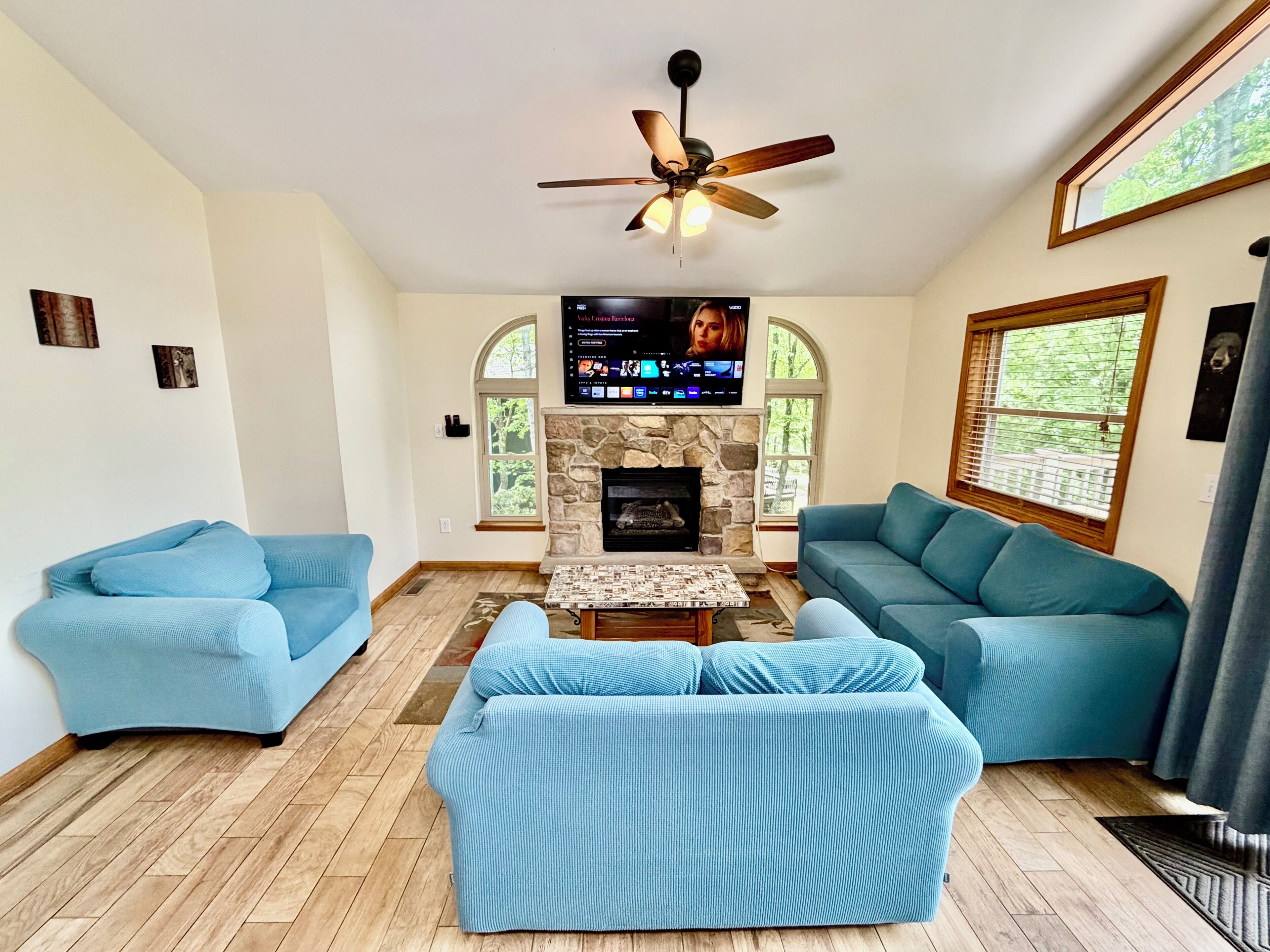 A living room with two blue sofas, a stone fireplace, a ceiling fan, and a TV mounted above the fireplace.