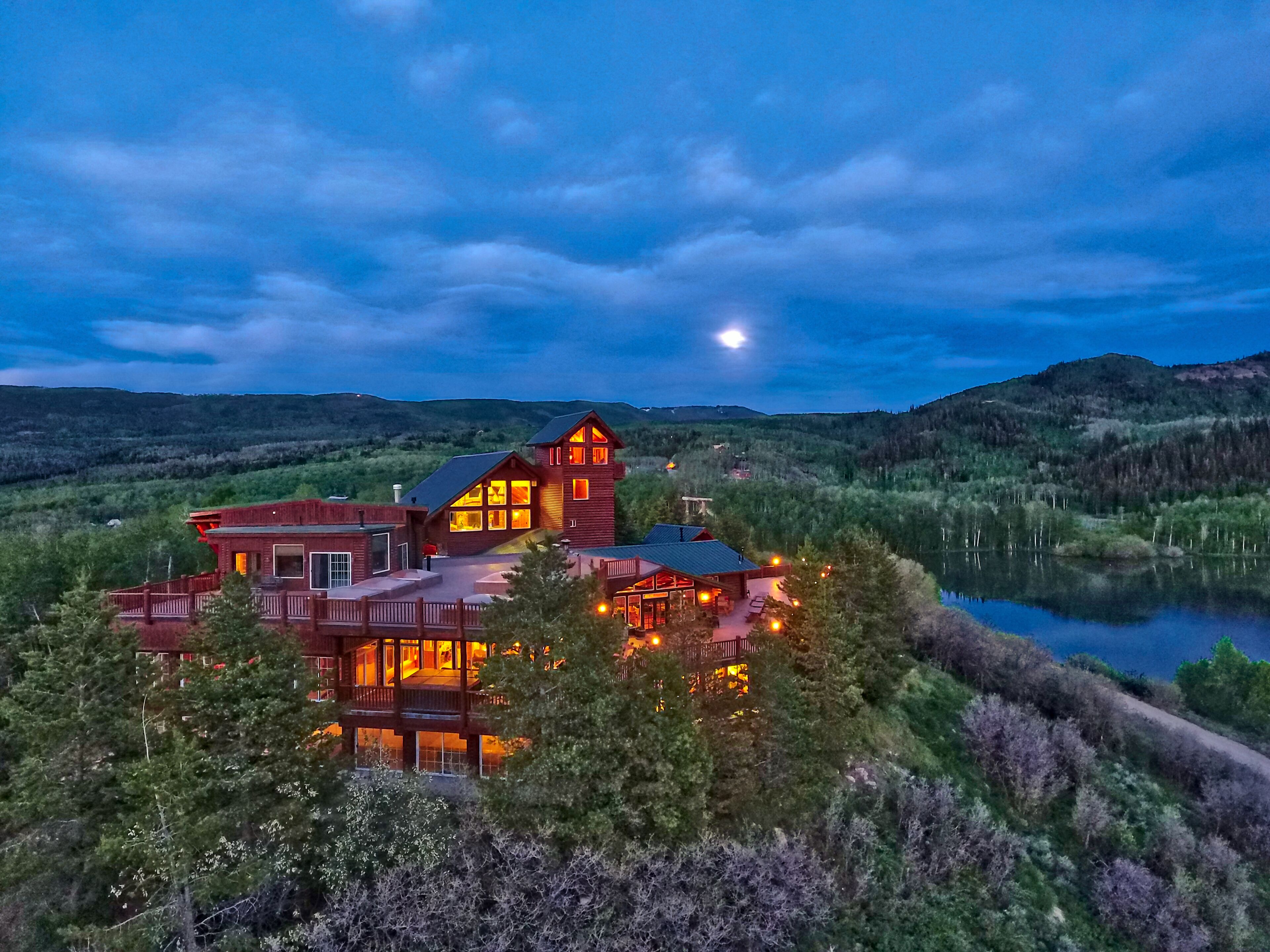A large, multi-story lodge with a prominent tower, situated on a lakeside with surrounding forested hills.