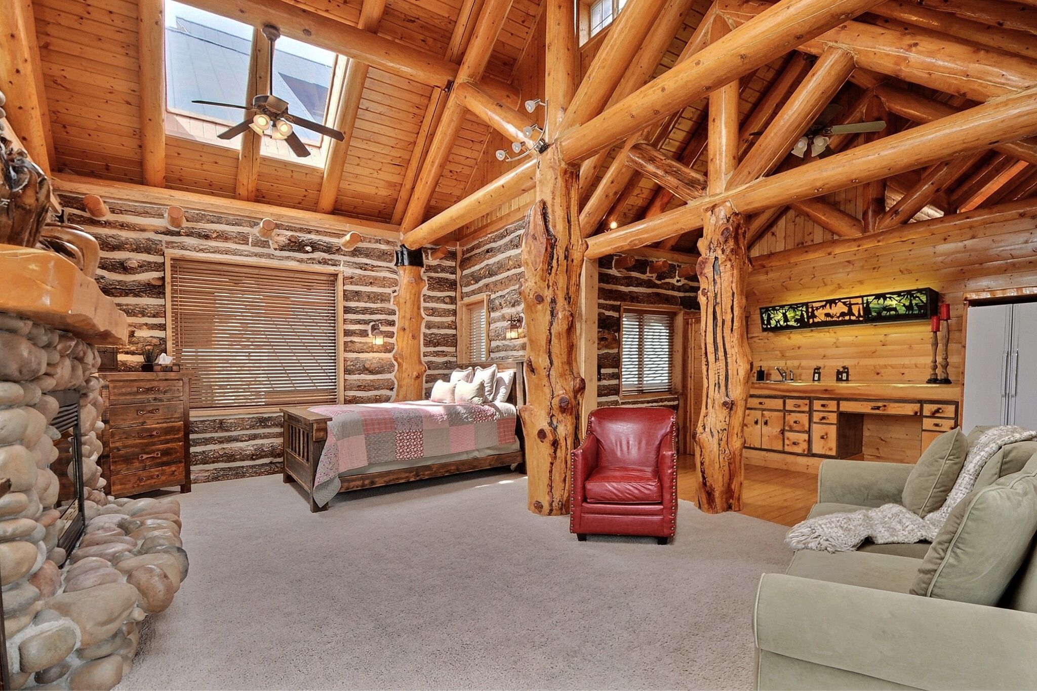 A log cabin interior with a bed, a red leather chair, a green sofa, a stone wall, and a wooden desk with a computer.