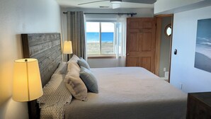 A bedroom with a bed, a ceiling fan, a window with curtains, and a view of the beach.