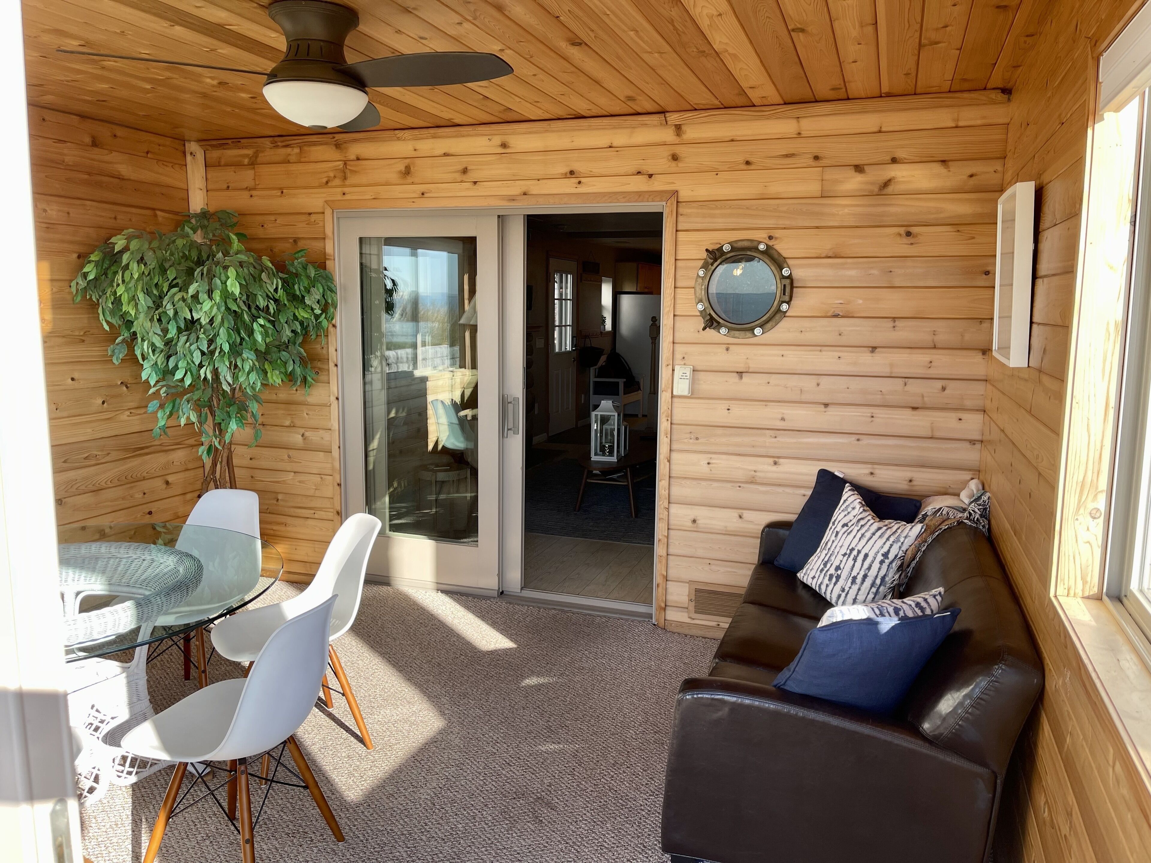 Un accogliente interno di una baita con soffitto in legno, una ventola a soffitto, una porta vetrata che dà su un balcone, un tavolo da pranzo con sedie, un divano con cuscini e una pianta in vaso.