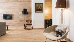 Un salón moderno con suelo de madera, un televisor de pantalla plana en la pared, una lámpara de pie negra, una silla de madera, un sillón blanco mullido y un cuadro de flores en la pared.