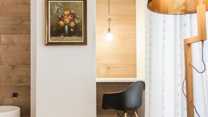 Una habitación moderna con piso de madera, una silla negra, un escritorio blanco, una lámpara colgante y un cuadro en la pared.