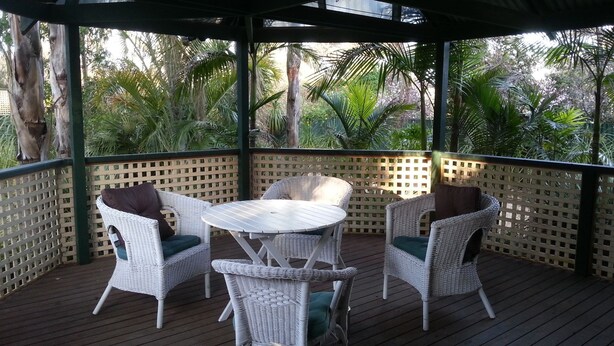 Un espace extérieur abrité, aménagé avec des meubles en osier et une table, entouré d’une terrasse en bois et d’une clôture à treillis.