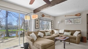 Ein Wohnzimmer mit einem Ecksofa, einem Couchtisch und einem großen Fenster mit Blick auf ein Haus und einen Baum.