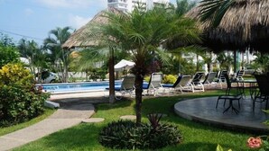 Un espace aménagé au bord de la piscine, avec des chaises longues, un palmier et une structure dotée d’un toit de chaume.