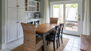 A dining area with a wooden table and wicker chairs, white cabinetry, and a view through a glass door to an outdoor patio. - Quintessential MV home near Oak Bluffs, beach & harbor (Oak Bluffs)