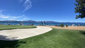 Campo de golf con un bunker de arena, un putting green y vista a un lago con barcos y montañas al fondo.