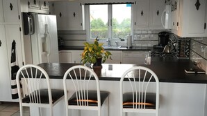 A modern kitchen with white cabinetry, a central island with two white chairs, a potted plant, and a window overlooking greenery. - Gorgeous Waterfront Home with Spectacular Views! Book Your Getaway Now!  (Lavallette)
