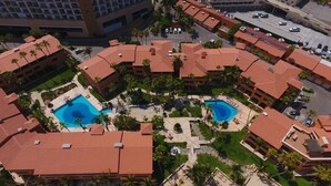 Eine Vogelperspektive zeigt ein Resort mit mehreren Gebäuden, zwei Schwimmbecken und Palmen.