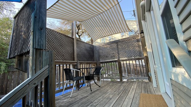 Une terrasse en bois avec un espace salon couvert, un paillasson et une vue sur un bâtiment en arrière-plan.