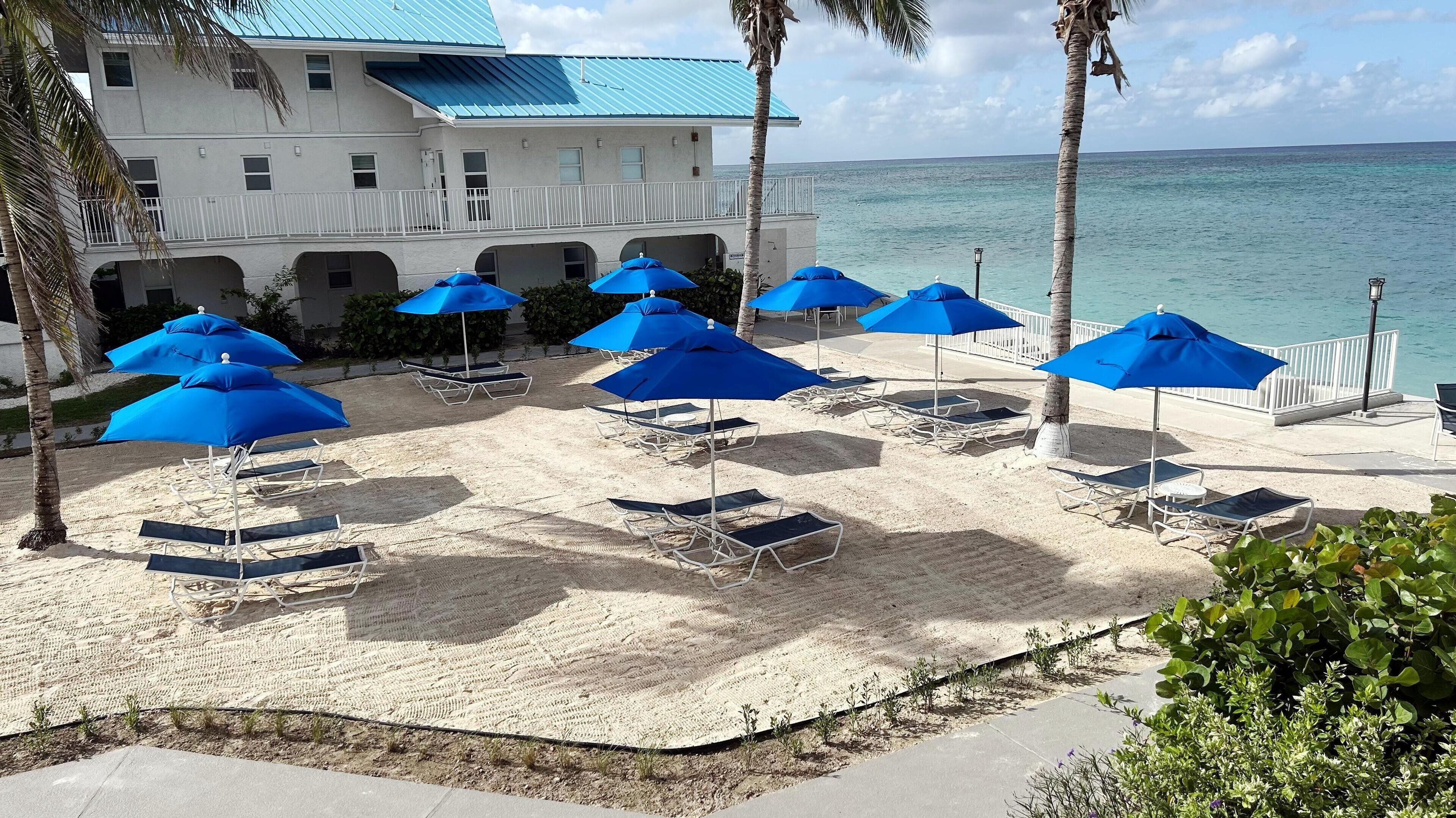 A beachside hotel with multiple blue umbrellas and lounge chairs, a white building with a blue roof, and a view of the ocean.