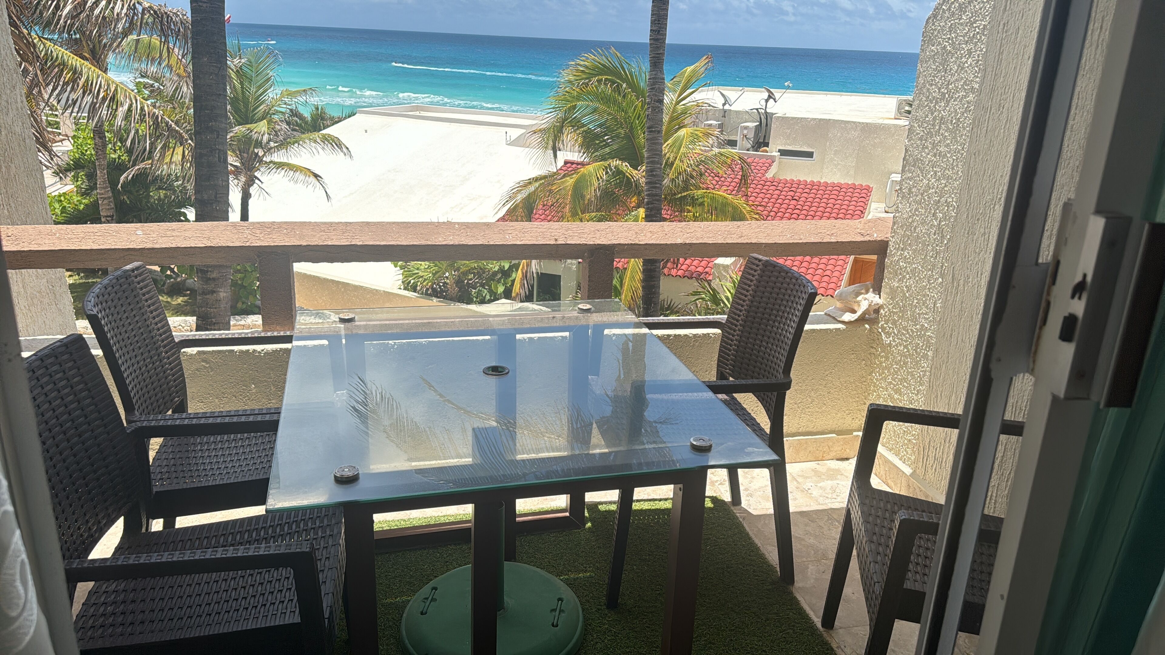 Un balcón con mesa y sillas de vidrio, con vista a una playa con palmeras y un cielo azul despejado.