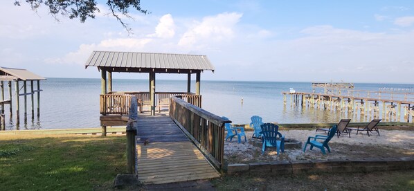 Eine Holzstege führt zu einem überdachten Sitzbereich mit blauen Stühlen und Blick auf das Wasser.