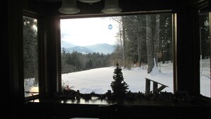 Habitación con ventana grande que ofrece vista a un paisaje nevado, un pequeño árbol de Navidad y un banco afuera.