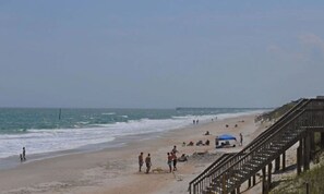 Ein Strand mit einer Holzpromenade, einem Pier in der Ferne und Menschen, die das Meer genießen.