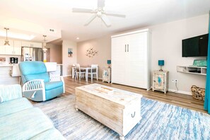 Un salon avec un canapé bleu, une table basse en bois, un coin repas avec des chaises blanches, une armoire blanche et un ventilateur de plafond.