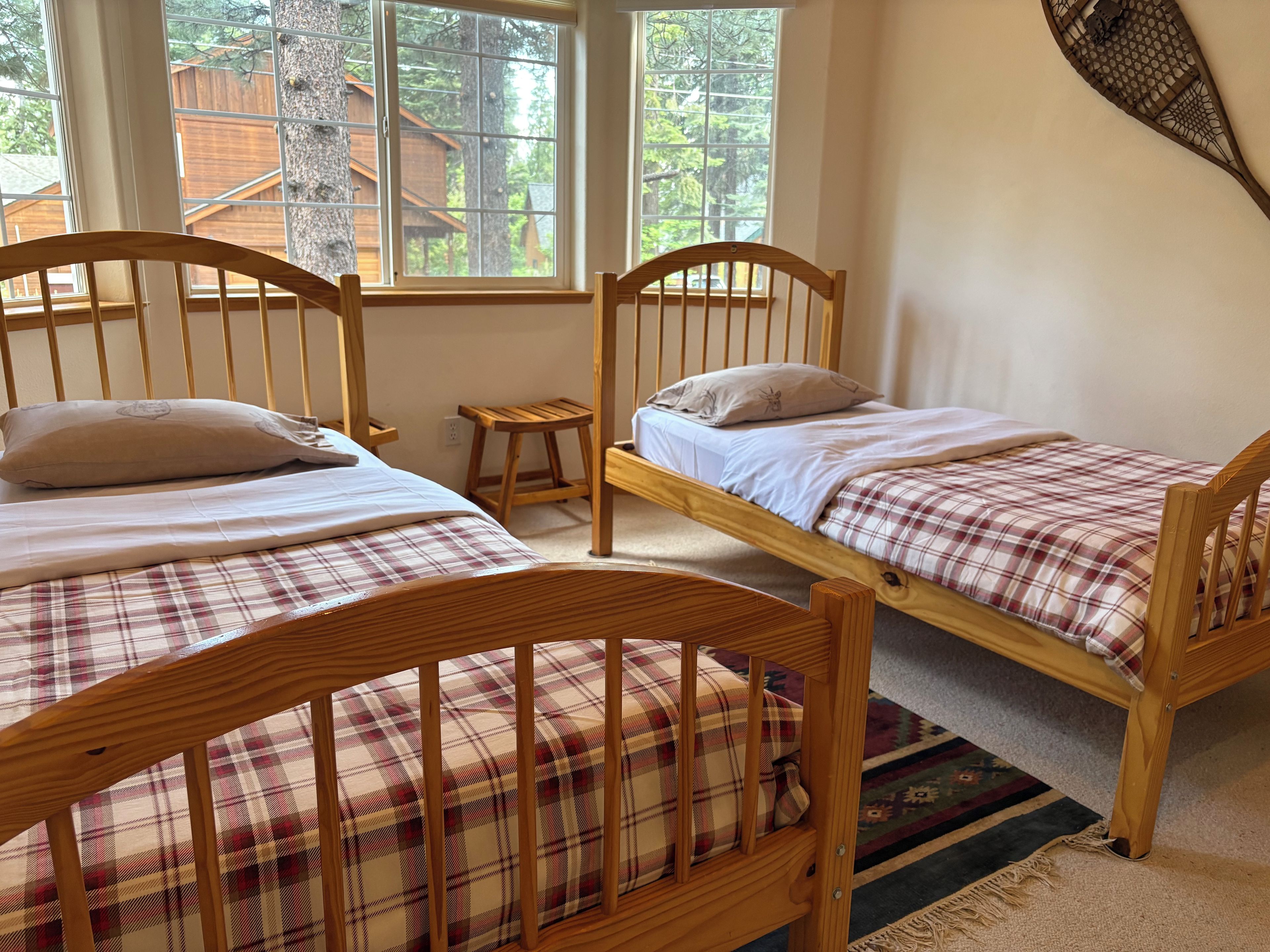 Una habitación con dos literas de madera, cada una con una almohada y una manta a cuadros. En el suelo, hay un taburete de madera y una alfombra colorida.