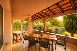 An outdoor patio with a wooden table and chairs, stone flooring, and a view of a garden with trees and a stone wall. - Holiday Apartment "Nikolina 1" with spacious garden (Rakalj)