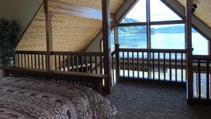 A bedroom with a sloped ceiling, large windows, a wooden railing, and a bed with patterned bedding.