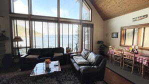 A cozy living room with a large window overlooking a body of water, a leather sofa, a coffee table with a vase of flowers, and a dining area with a checkered tablecloth.