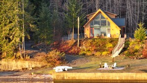 A lakeside cabin with a large glass window, a staircase leading to the main entrance, and a dock with chairs.