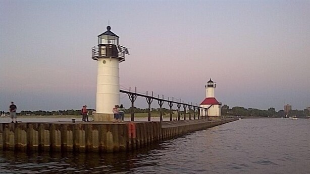 Un faro con la parte superior blanca y roja, ubicado al final de un muelle que se adentra en aguas tranquilas.