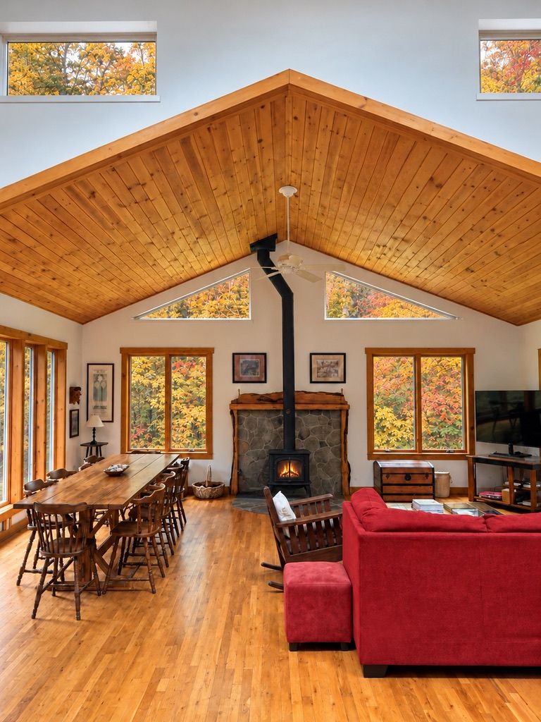 Ein geräumiges Wohnzimmer mit hoher Holzbalkendecke, Kamin, Essbereich mit Holzmöbeln und großen Fenstern mit Herbstlaub.