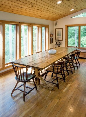 Une longue table à manger en bois, avec six chaises, de grandes fenêtres et un plafond en bois.