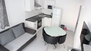 A compact kitchen with white cabinets, a microwave, a stove, and a sink. There is a small dining area with a glass table and two chairs. A gray sofa is positioned in the living area. - Danica - apartment 003 (Promajna)