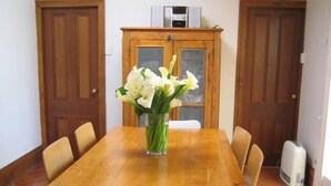 A wooden dining table with a vase of white flowers, surrounded by six chairs, in a room with wooden doors and a tiled floor.