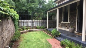 A backyard with a grassy lawn, a stone wall, a wooden fence, and a covered patio area with potted plants.