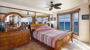 A bedroom with a wooden bed, a ceiling fan, a large mirror, and a view of the ocean. - Absolutely Stunning Oceanfront Condo! (Koloa)