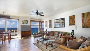 A spacious living area with a large window overlooking the ocean, a ceiling fan, and a variety of seating options including a sofa and chairs. - Absolutely Stunning Oceanfront Condo! (Koloa)