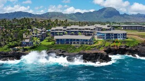 A coastal hotel with multiple buildings, a rocky shoreline, and a mountainous backdrop. - Absolutely Stunning Oceanfront Condo! (Koloa)
