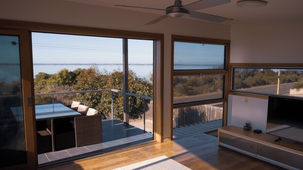 A modern interior with large windows, a ceiling fan, and a balcony with outdoor furniture. - Camelot - opposite the foreshore. Stunning sea views. (Kingscote)