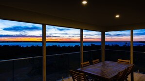 Un balcon avec une table et des chaises en bois, de grandes fenêtres offrant une vue sur un coucher de soleil sur l  ’eau.