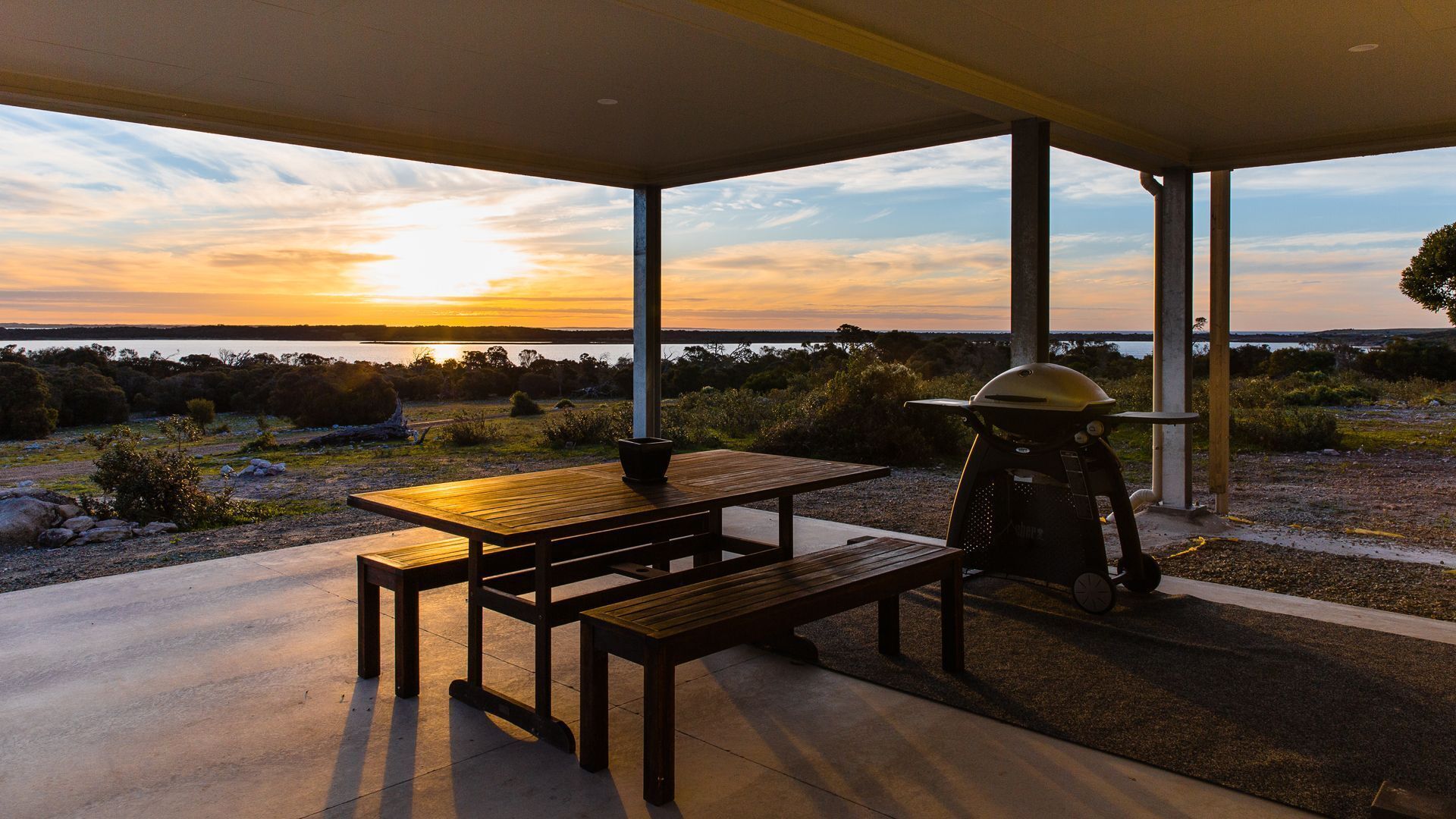 A covered outdoor area with a picnic table and a grill, overlooking a scenic landscape with a sunset.