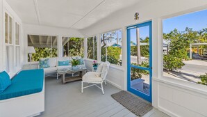 A covered porch with white furniture, including a sofa and chairs, and a view of the street through large windows.