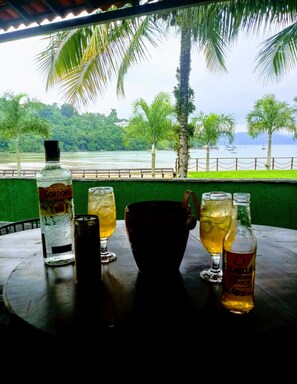 A table with drinks and a bottle, overlooking a beach with palm trees and boats.