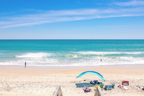 A beach with a clear sky, waves, and a person walking. There are beach chairs and a blue umbrella.