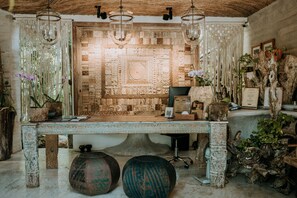 A room with a large, intricately carved stone wall, a wooden ceiling, and hanging light fixtures. There are potted plants and a desk with a chair.