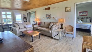 A cozy living room with a wooden ceiling, a beige sofa, a coffee table, and a wicker chair. There is a bed in the adjacent room with a striped bedspread. - Great Gulf Views and easy walk to the beach.  Dog Friendly! (Gulf Shores)